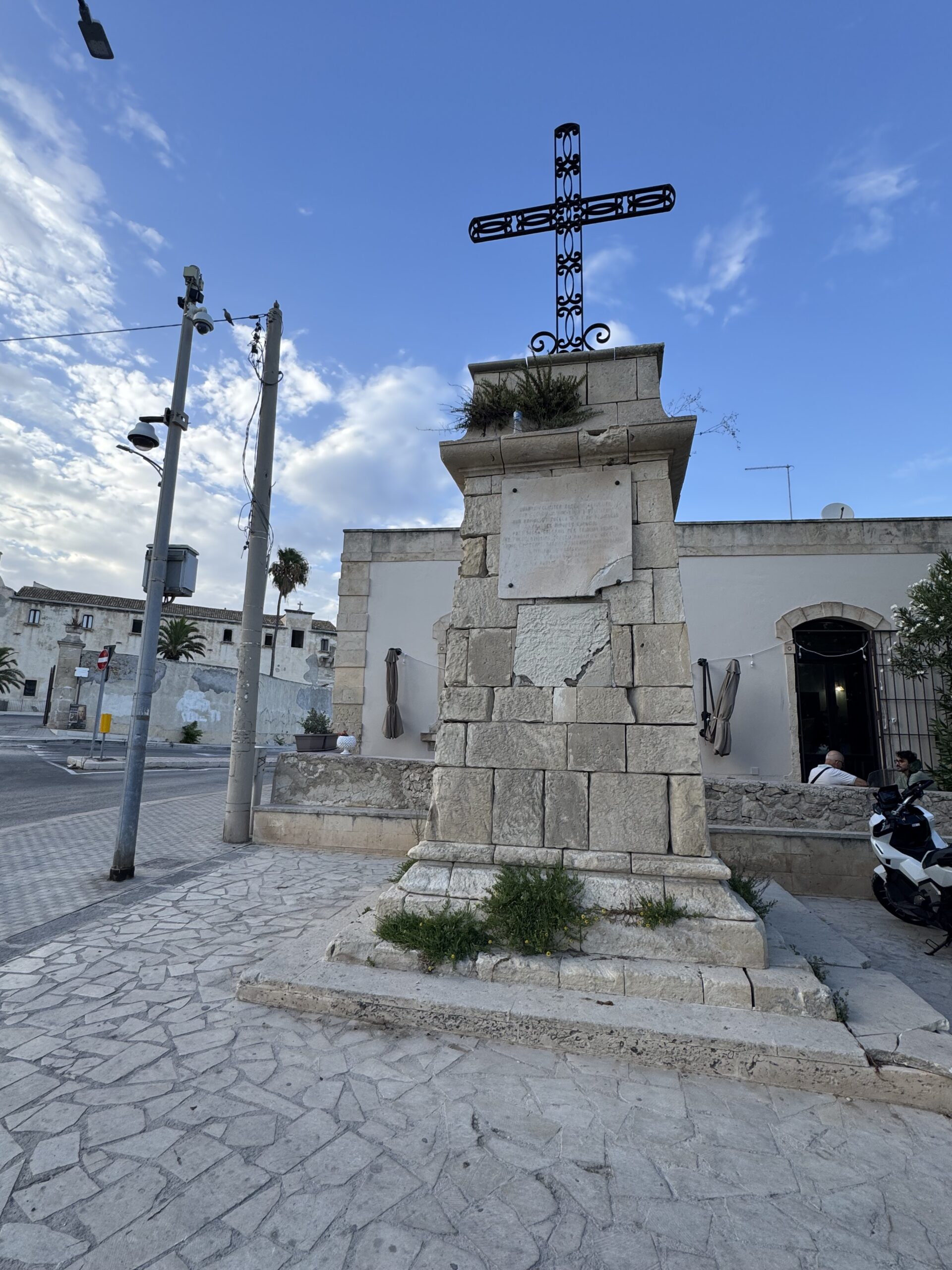 La Croce delle Sante Missioni ai Cappuccini vista da piazza Cappuccini, con il basamento in pietra e la croce in ferro, sullo sfondo la strada e gli edifici.