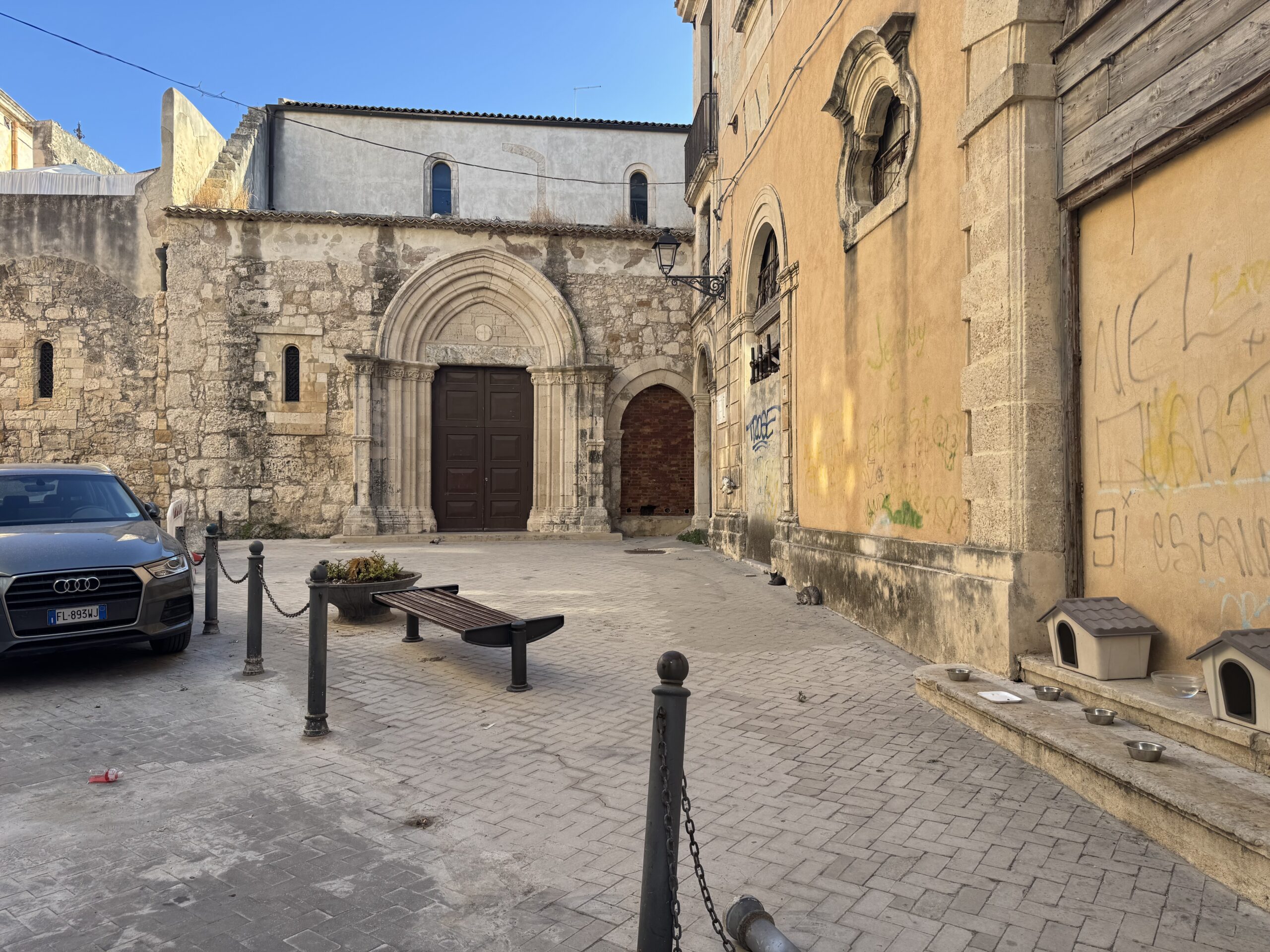 Veduta della chiesa di San Pietro Apostolo in Ortigia con la piazzetta pavimentata, una panchina centrale e alcune auto parcheggiate attorno.