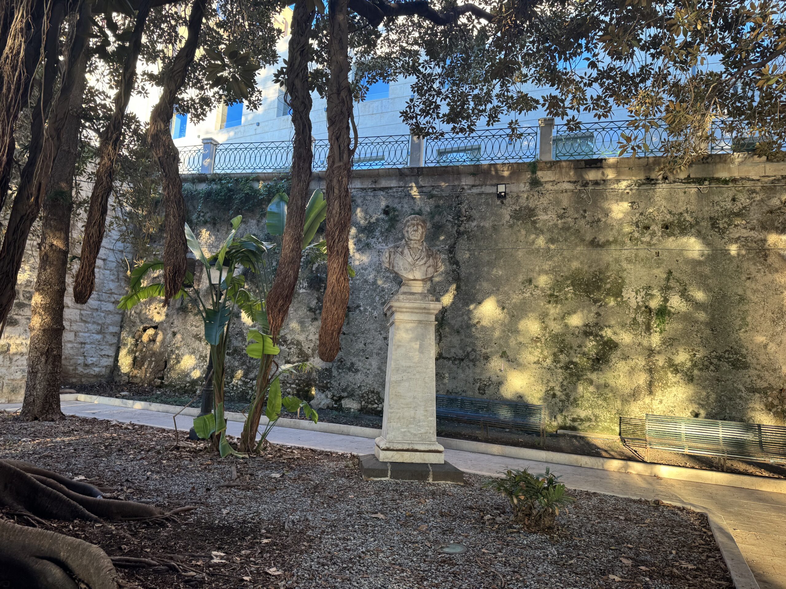 Veduta ampia del busto marmoreo di Carmelo Campisi su alto piedistallo nella Villetta Aretusa a Siracusa, con alberi in primo piano e panchine lungo il viale.