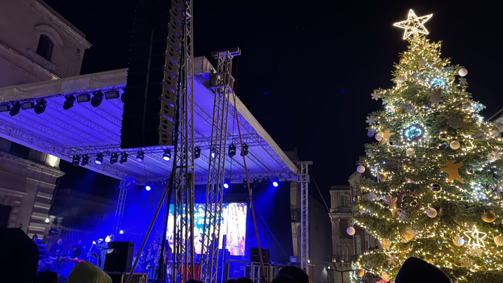 Palco del concerto con luci blu e maxi schermo in Piazza Duomo a Siracusa, con grande albero di Natale illuminato, pochi minuti prima della mezzanotte.