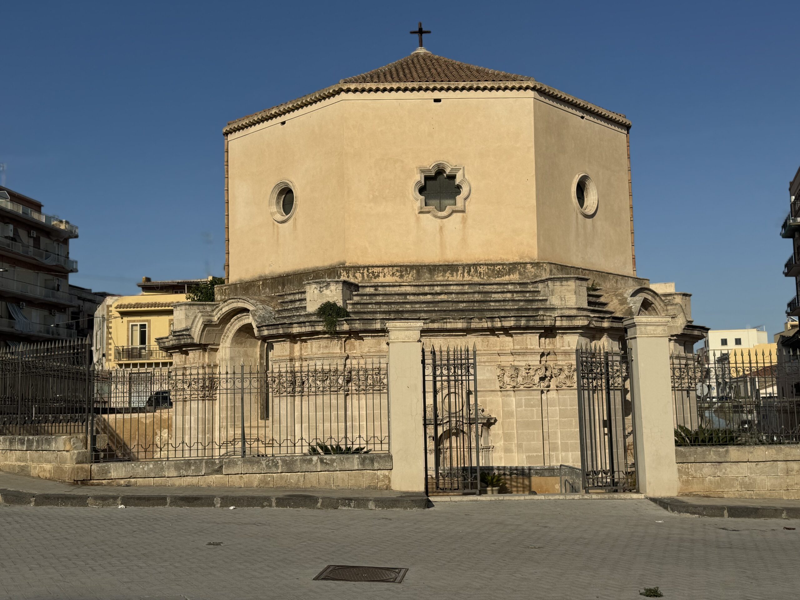 Vista esterna del tempietto ottagonale della Chiesa di Santa Lucia al Sepolcro a Siracusa, con tetto a coppi e croce, finestre circolari e finestra quadrilobata, recinzione in ferro.