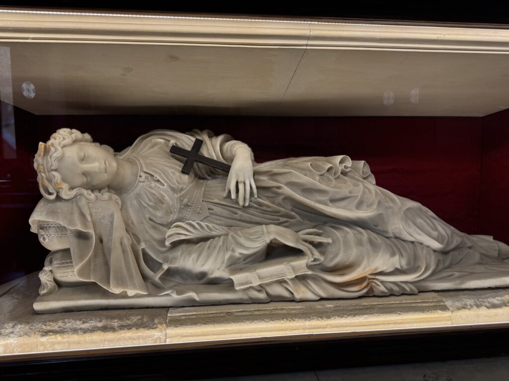 Statua marmorea della “Santa Lucia dormiente” di Gregorio Tedeschi in teca, con croce sul petto e libro ai piedi, fondo rosso.
