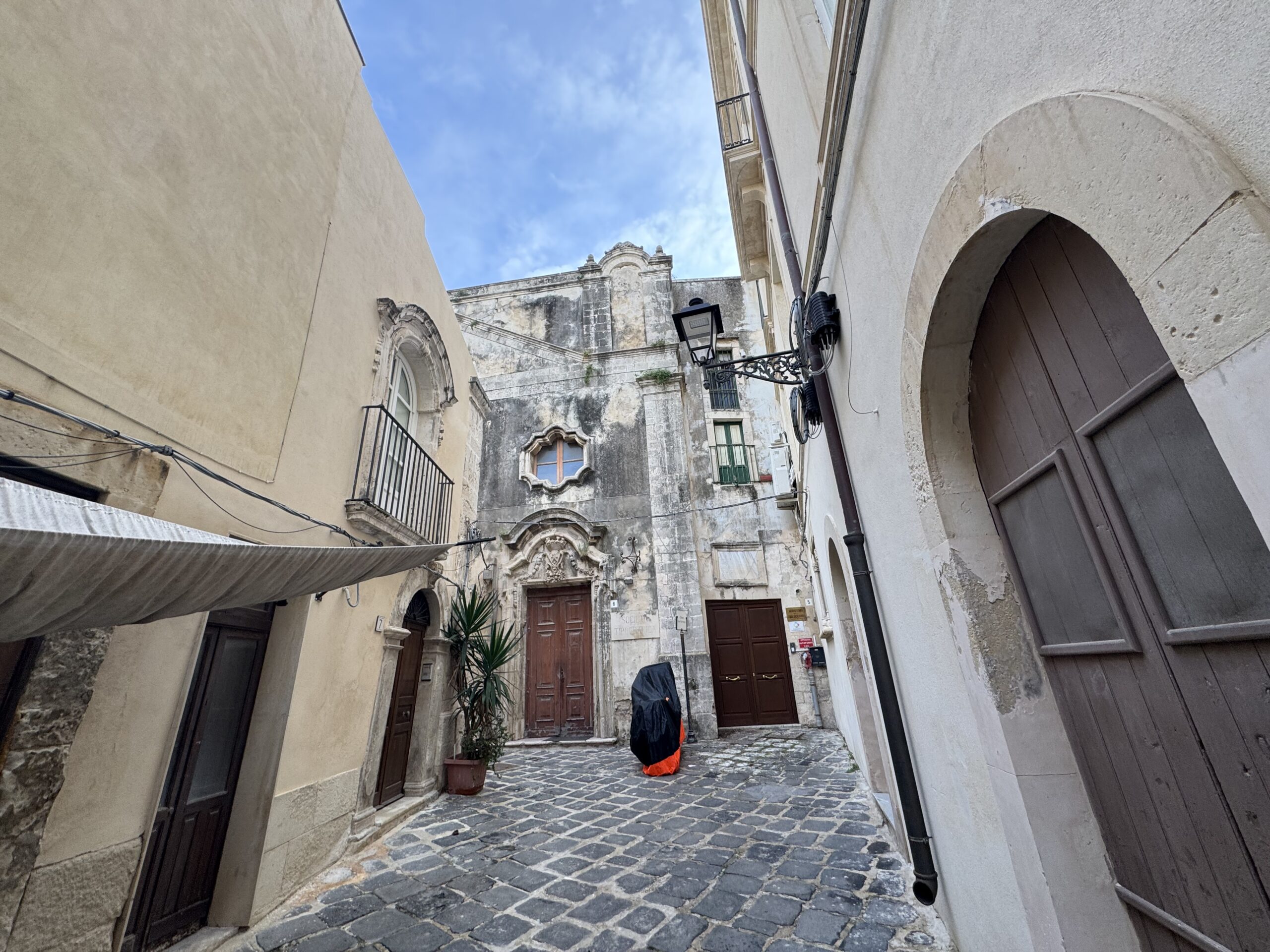 Vicolo lastricato che termina su un piccolo slargo; in fondo la facciata in pietra dell’ex chiesa di San Salvatore con portale barocco, porta lignea e finestra polilobata; scooter coperto al centro.