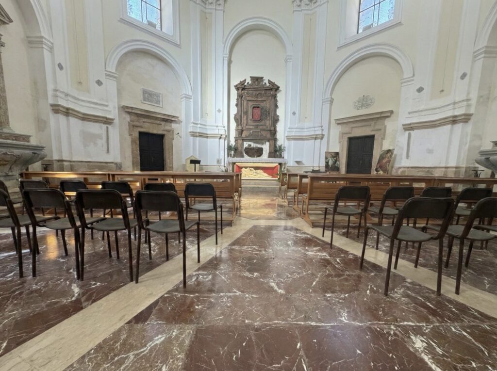 Interno del Tempietto di Santa Lucia al Sepolcro con sedie, pavimento in marmo e altare sul fondo con teca illuminata.