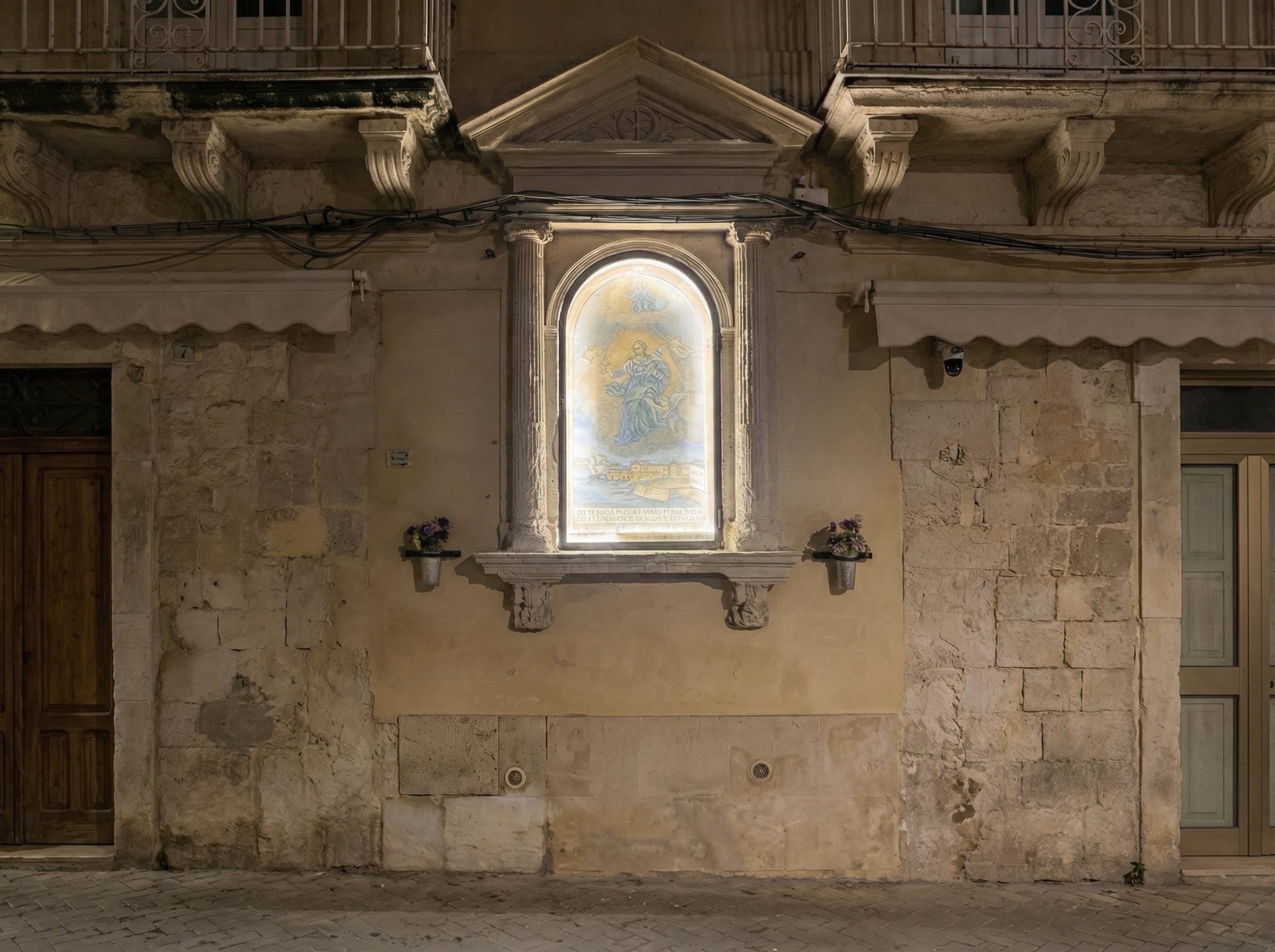 Edicola votiva di Santa Lucia incassata in una facciata in pietra, con nicchia ad arco illuminata e immagine della Santa; ai lati due colonne scanalate e due portafiori.8