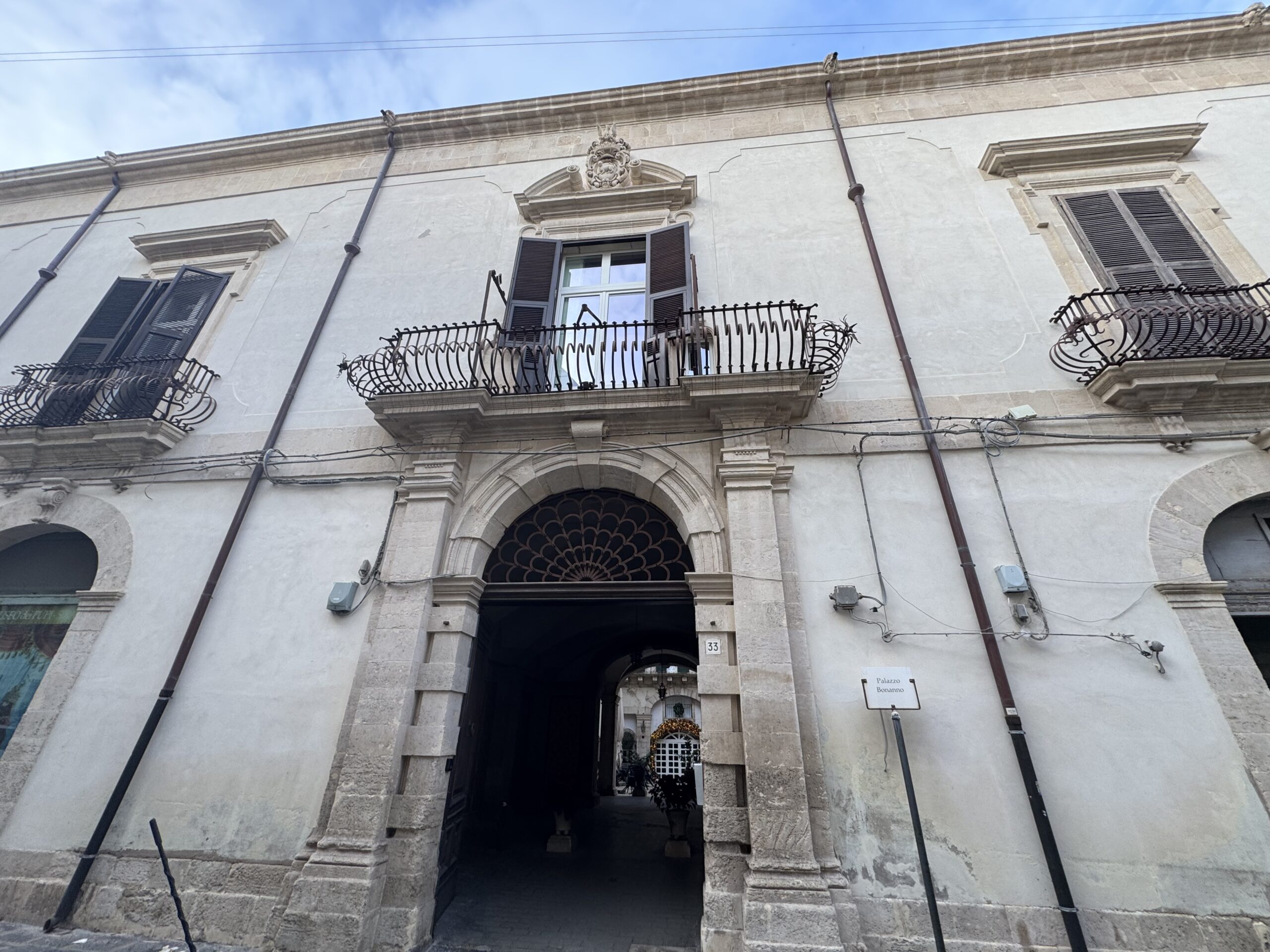 Facciata di Palazzo Bonanno in via della Maestranza a Ortigia: portale arcuato con bugne, balcone con inferriata bombata in ferro battuto e stemma araldico della famiglia Bonanno nel timpano spezzato