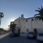 Veduta frontale della facciata della chiesa di Santa Maria della Misericordia e dei Pericoli a Siracusa, illuminata dalla luce calda del tramonto, con palme e auto in primo piano sfocate per privacy