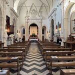 Vista interna della navata unica della chiesa, con la pavimentazione bianca e nera a chevron, le panche in legno e l'altare maggiore in fondo, sormontato da arcate sostenute da colonne con timpano spezzato