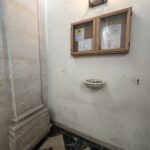 Acquasantiera in marmo bianco a forma di conchiglia fissata alla parete vicino all'ingresso della chiesa, accanto alla bacheca degli avvisi parrocchiali