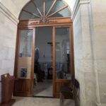Porta a doppio battente in legno con sopraluce semicircolare a raggiera in vetro, attraverso la quale si intravede l'interno di una cappellina con statua, crocifisso e altare laterale