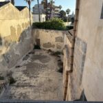 Vista dall'alto di un cortile esterno o tratto di antico fossato che corre lungo la parete del convento, con muri scrostati e una grondaia in pietra; sullo sfondo edifici moderni e palme