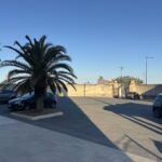Veduta della piazza dei Cappuccini con il muro di cinta del complesso conventuale, due alte palme su un'aiola, le cancellate di accesso al cortile e auto in lontananza