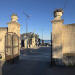 Cancello in ferro battuto di ingresso alla Latomia dei Cappuccini, fiancheggiato da due pilastri in pietra sormontati da grandi vasi con piante di agave; sullo sfondo la strada con vista sul mare
