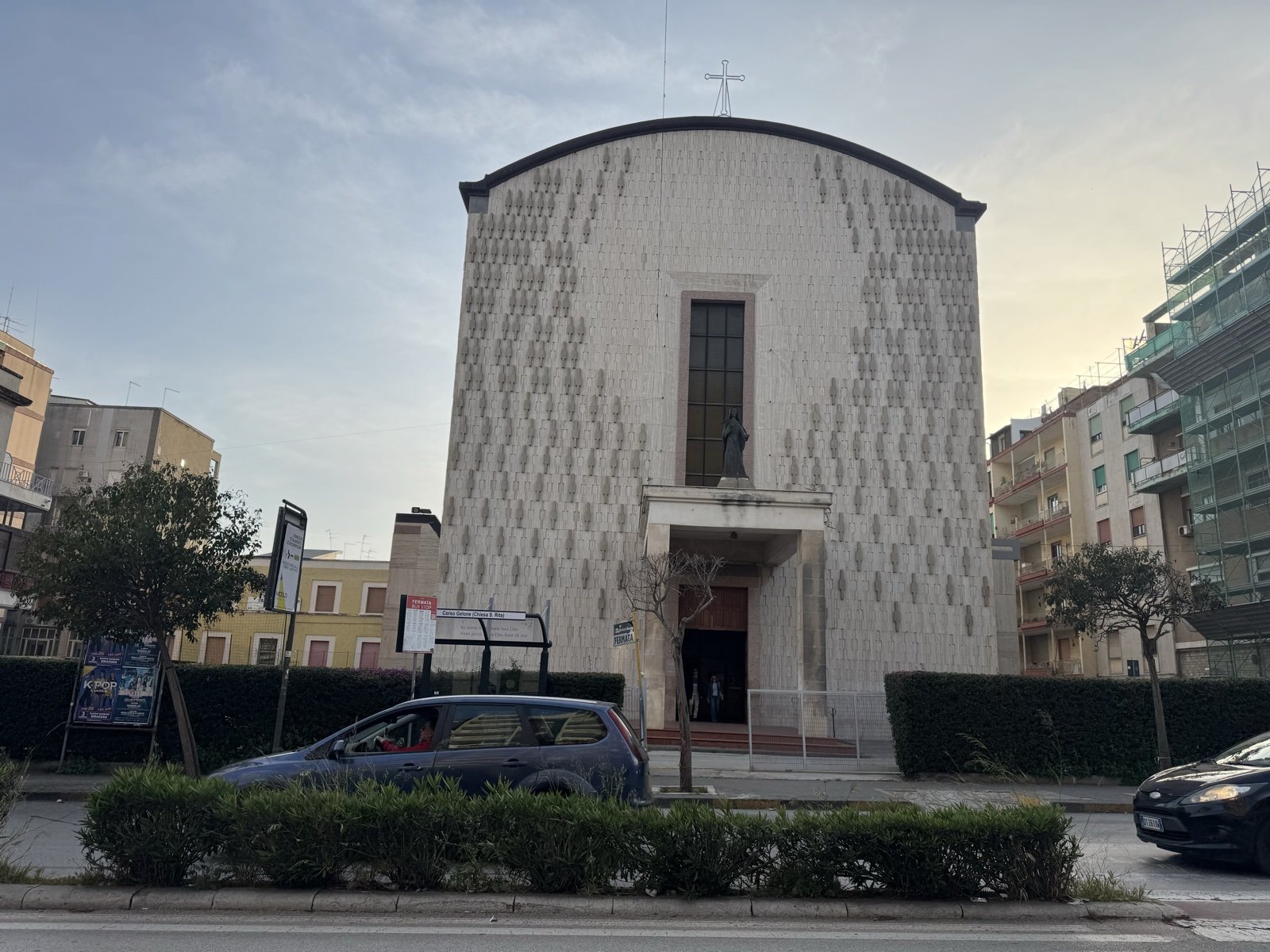 Facciata della Chiesa di Santa Rita da Cascia, Corso Gelone 93, Siracusa: fronte bianco a vela rivestito in lastre di pietra calcarea iblea con decorazione a foglie in rilievo, portale centrale sormontato da statua bronzea della santa e grande finestra verticale