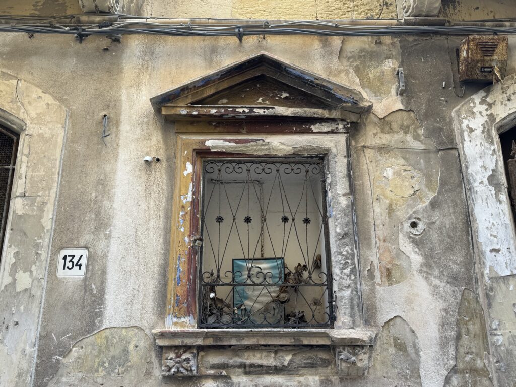 Edicola votiva vuota al civico 134 di Via Dione a Ortigia, con nicchia in pietra calcarea, timpano triangolare e grata in ferro battuto