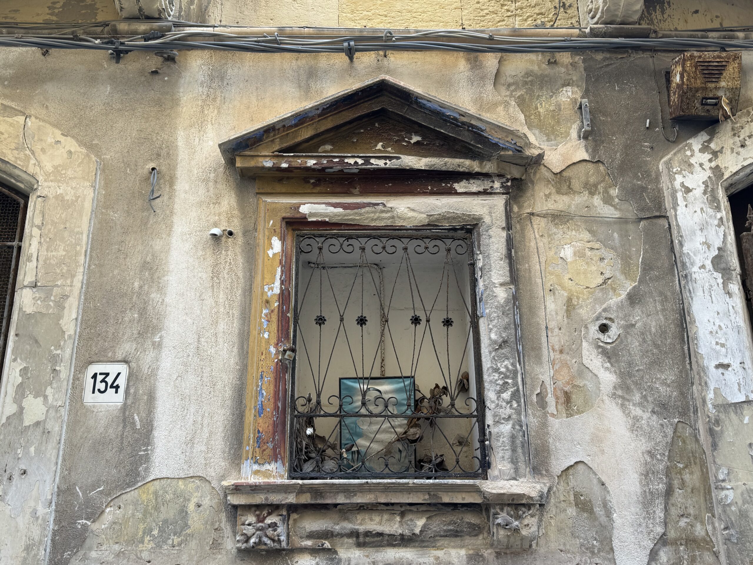 Edicola votiva vuota al civico 134 di Via Dione a Ortigia, con nicchia in pietra calcarea, timpano triangolare e grata in ferro battuto