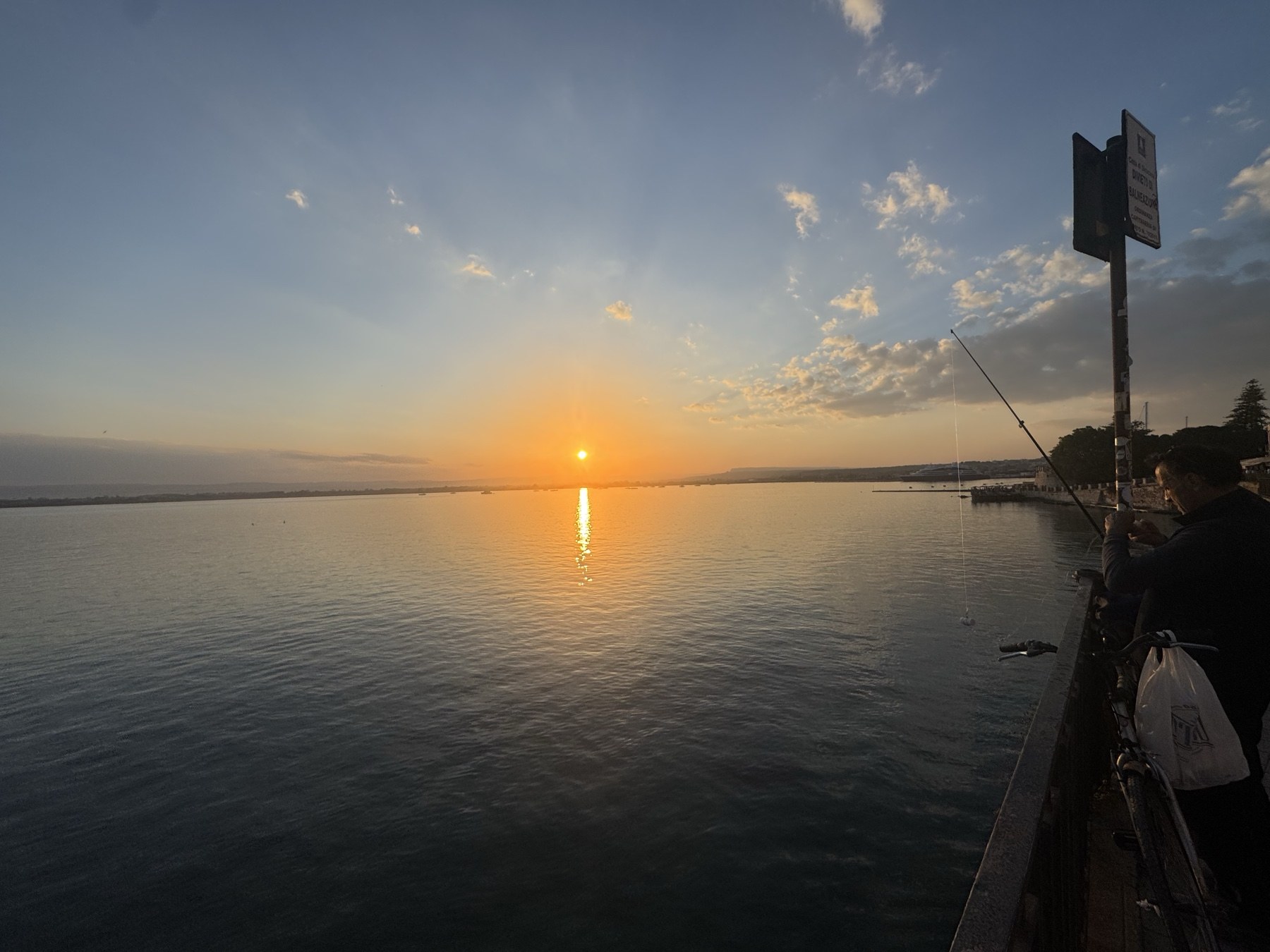 Pescatore con canna sul Lungomare Alfeo al tramonto, con asta dei segnali e cartello AST in primo piano