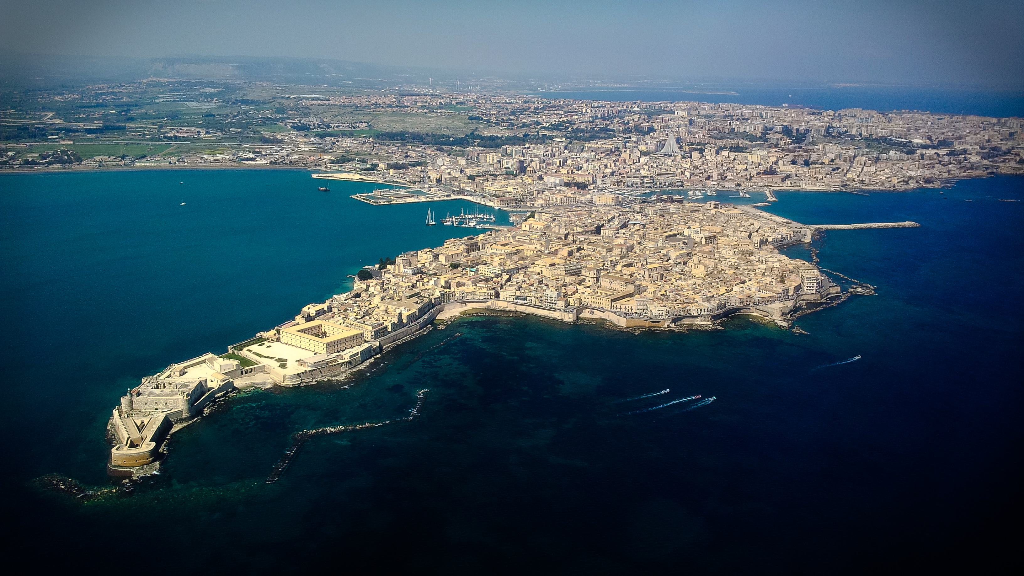 Veduta aerea dell'isola di Ortigia circondata dal mare, con il tessuto edilizio storico, la darsena e i ponti verso la terraferma