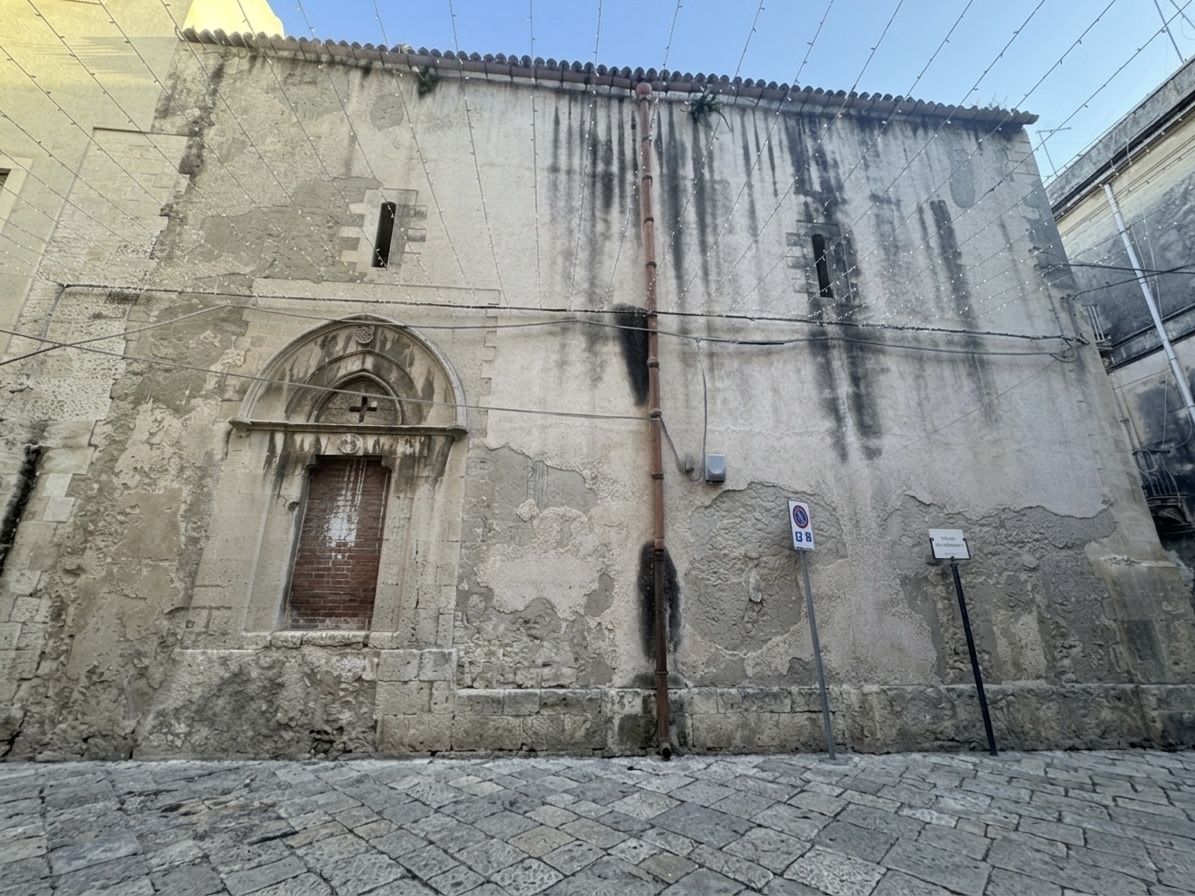 Prospetto laterale della Chiesa di San Tommaso: portale ogivale gotico murato con croce scolpita e due strette feritoie nella parte alta della parete.