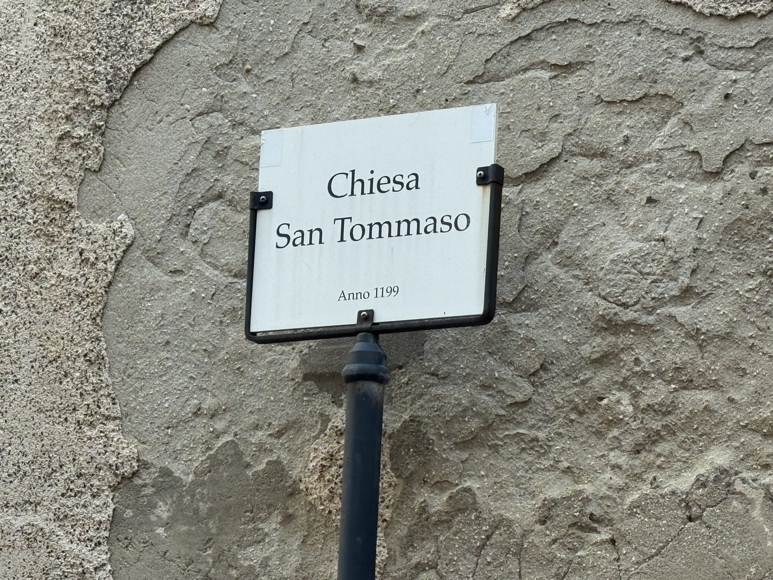 Targa rettangolare bianca con cornice nera con la scritta Chiesa San Tommaso Anno 1199.