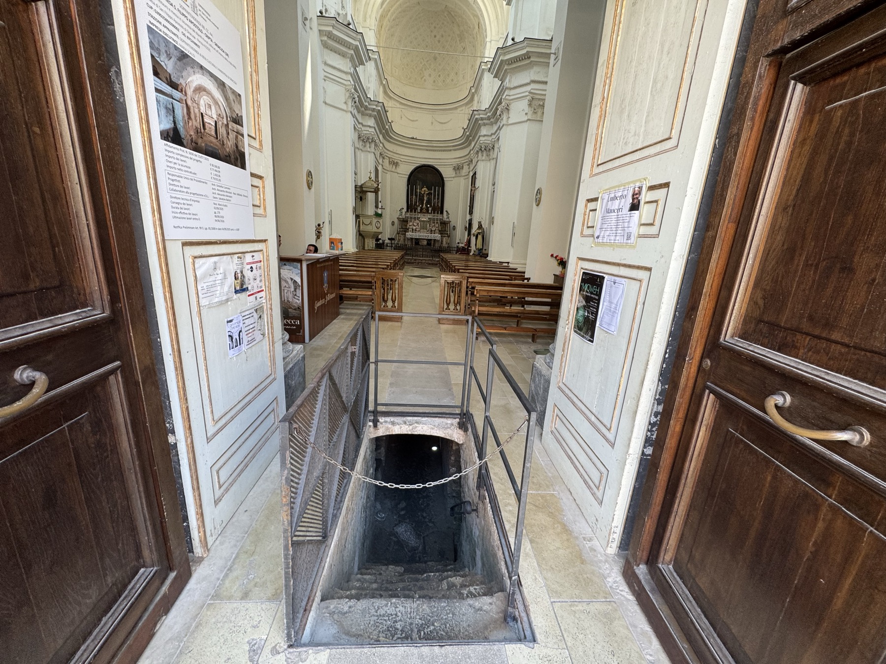 Botola con ringhiera metallica e scala in pietra che scende verso i sotterranei, all'interno della chiesa, con la navata e l'abside sullo sfondo.