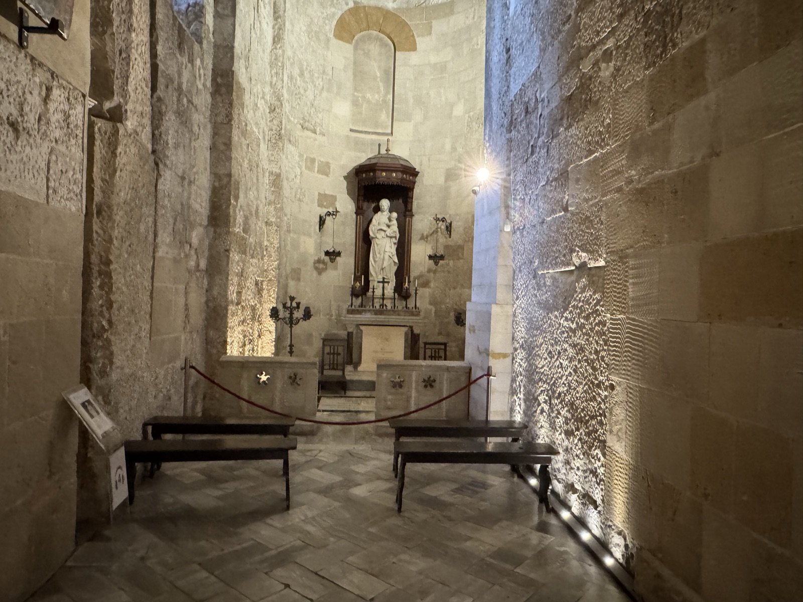 Vista d'insieme della Cappella della Madonna della Neve del Duomo di Siracusa con pareti in pietra a vista dell'apside bizantino-normanno superstite, statua marmorea di Antonello Gagini sotto baldacchino ligneo, candelabri laterali, panche e lampade votive