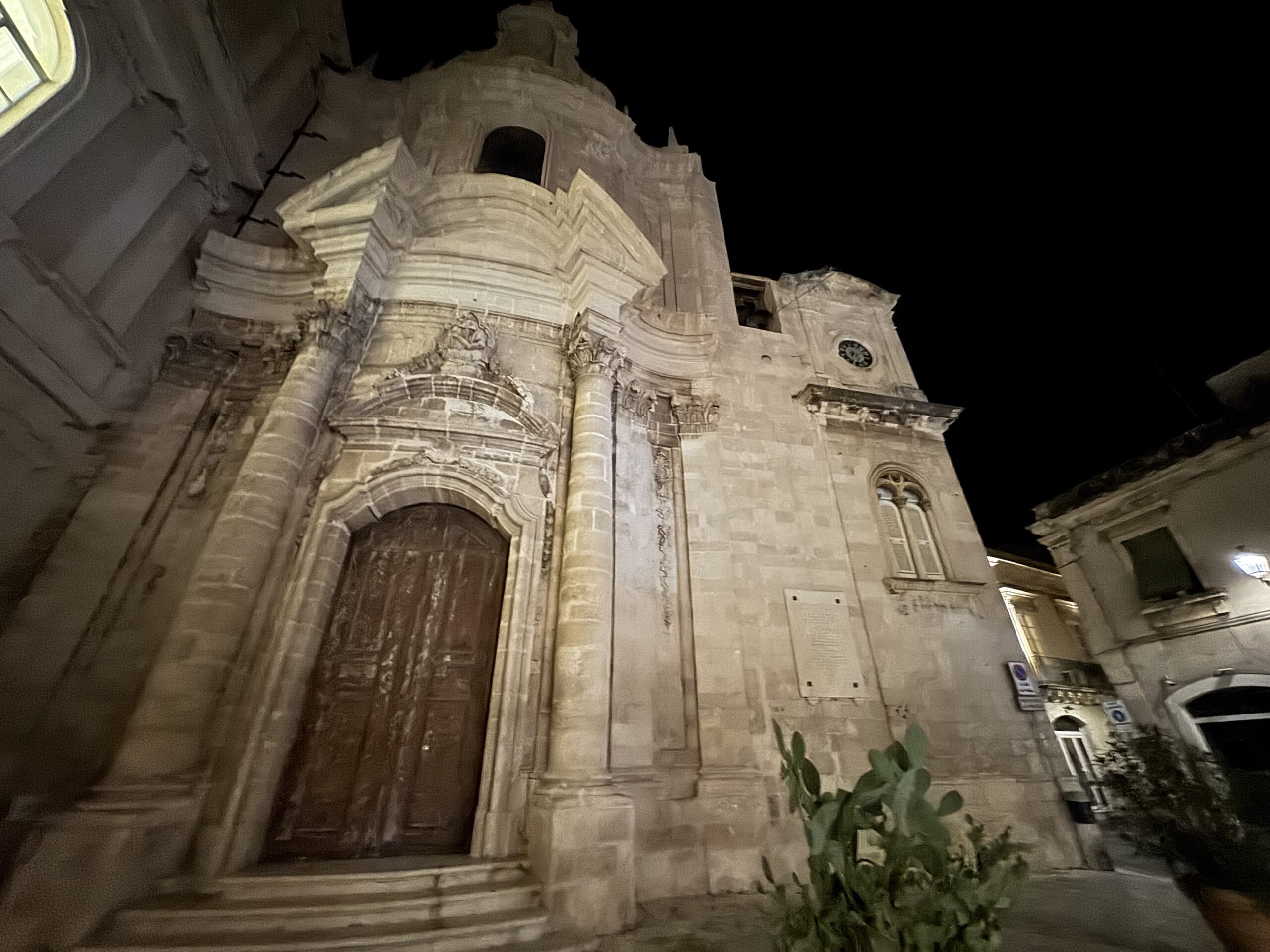 Facciata convessa tardo-barocca della chiesa illuminata di notte, con il campanile-orologio sulla destra
