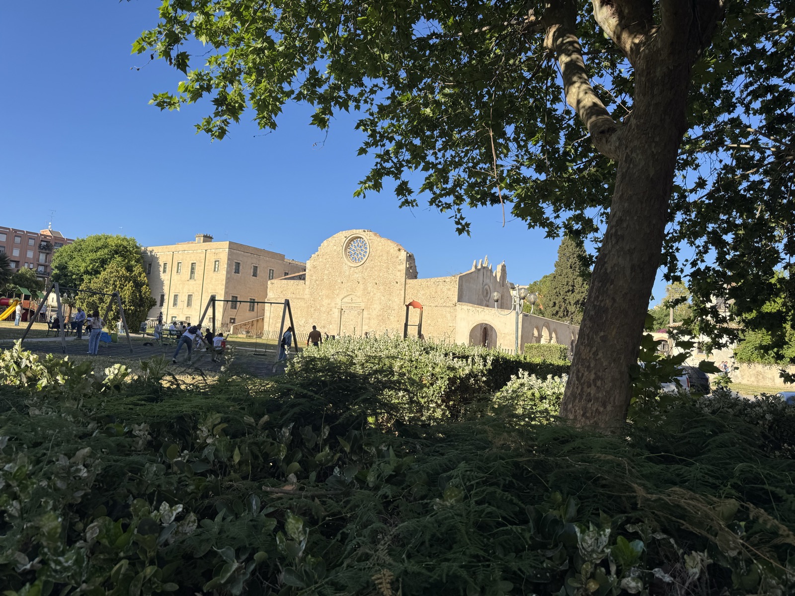 Chiesa di San Giovanni alle Catacombe vista dal Piazzale San Marziano: viale alberato in primo piano, siepi di vegetazione mediterranea, parcheggio antistante.