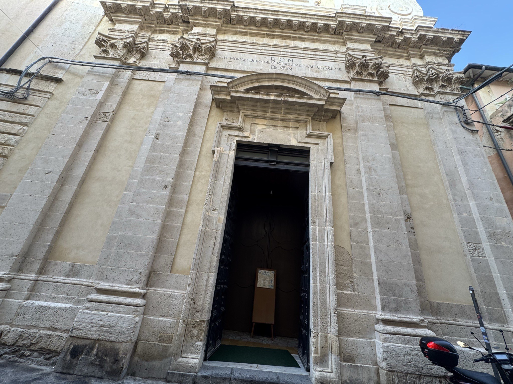Portale d'ingresso aperto della chiesa, con bussola lignea e cartello informativo all'interno; facciata in conci di pietra calcarea giallastra.