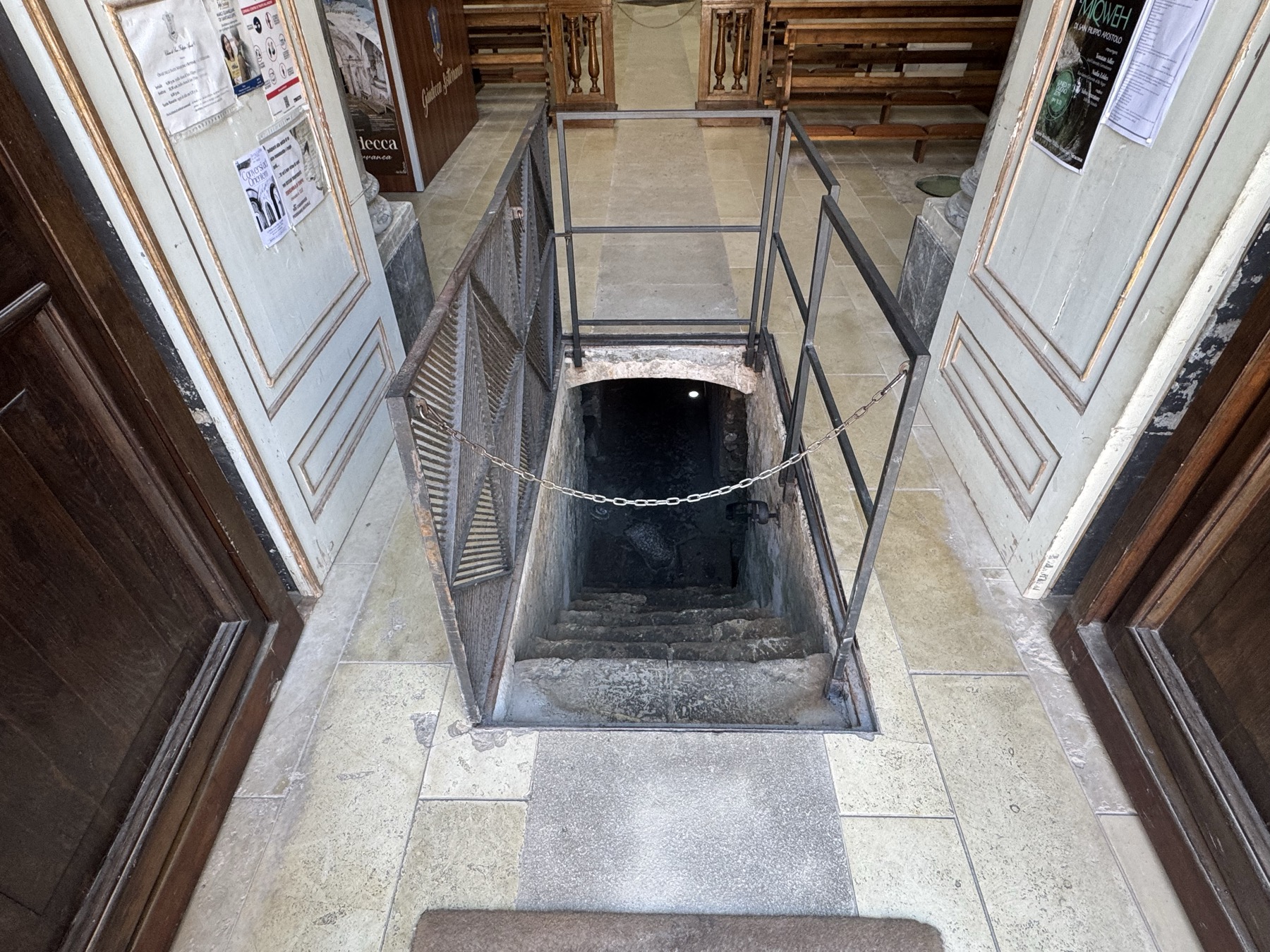 Stretta scala in pietra che scende nel buio dei sotterranei, delimitata da una ringhiera metallica con catena.