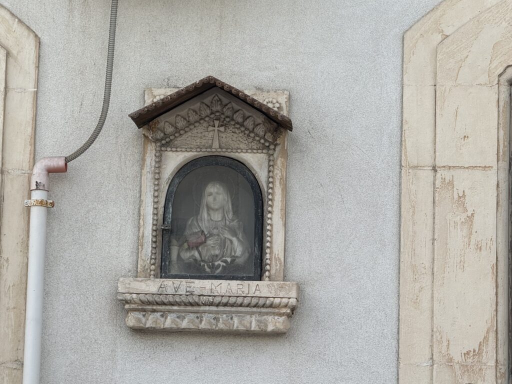 Dettaglio del frontone triangolare con croce greca e decorazione a rosario di perline, e iscrizione «Ave = Maria» sulla mensola sottostante