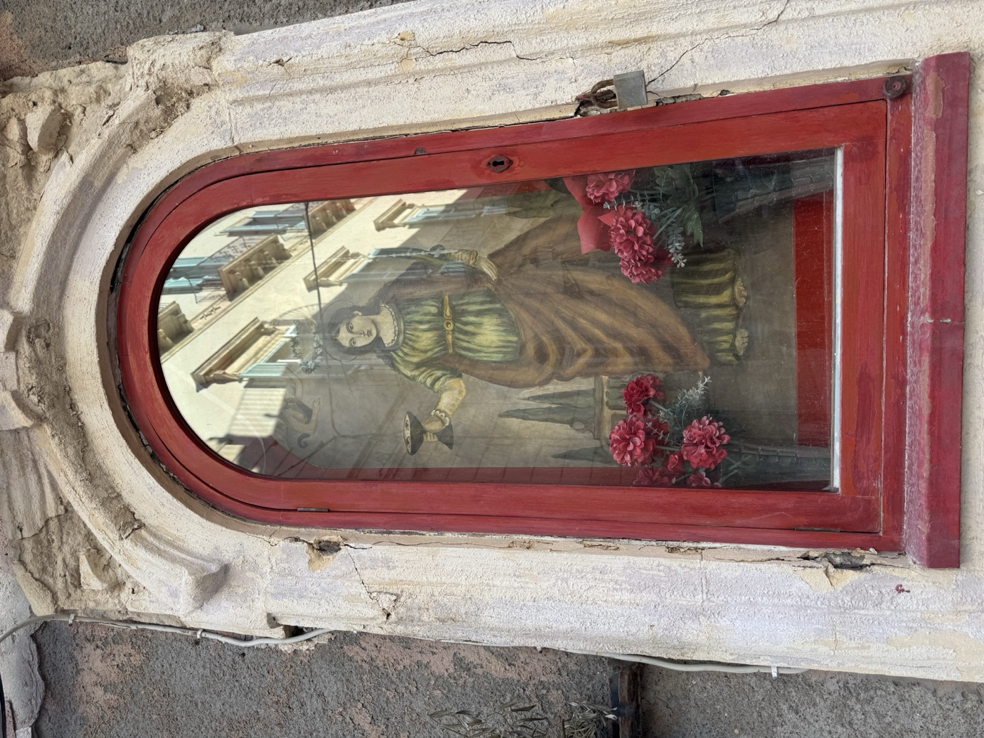 Dettaglio del dipinto popolare devozionale di Santa Lucia con tunica verde, mantello rosso, palma del martirio nella mano sinistra, piattino con gli occhi e fiamma divina nella destra, due putti che reggono la corona dorata