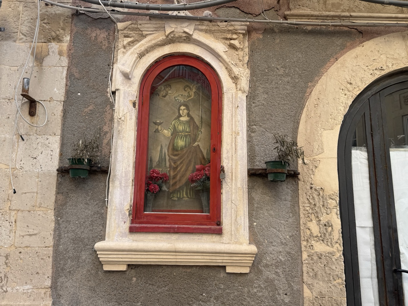 Edicola votiva di Santa Lucia in via della Maestranza 136, Ortigia, stato attuale, con cornice in pietra calcarea bianca aperta a orecchioni superiori, sportello in ferro battuto a raggiera, cornice interna lignea verniciata di rosso aggiunta dopo il 2010 e dipinto popolare della santa con piatto degli occhi, fiamma divina, palma del martirio e due putti incoronanti