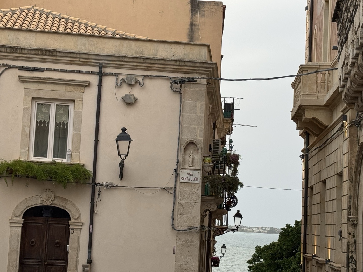 Vista urbana del cantonale fra via Pompeo Picherali e piazzetta San Rocco Ortigia con edicola di Santa Lucia, palazzi storici e mare del Porto Grande di Siracusa sullo sfondo