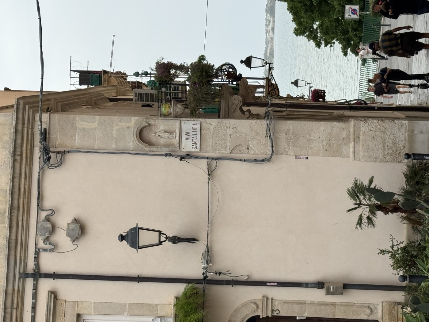 Vista panoramica della piazzetta San Rocco a Ortigia con edicola di Santa Lucia sul cantonale, cornicione modanato in pietra calcarea del palazzo e persone che attraversano la piazzetta