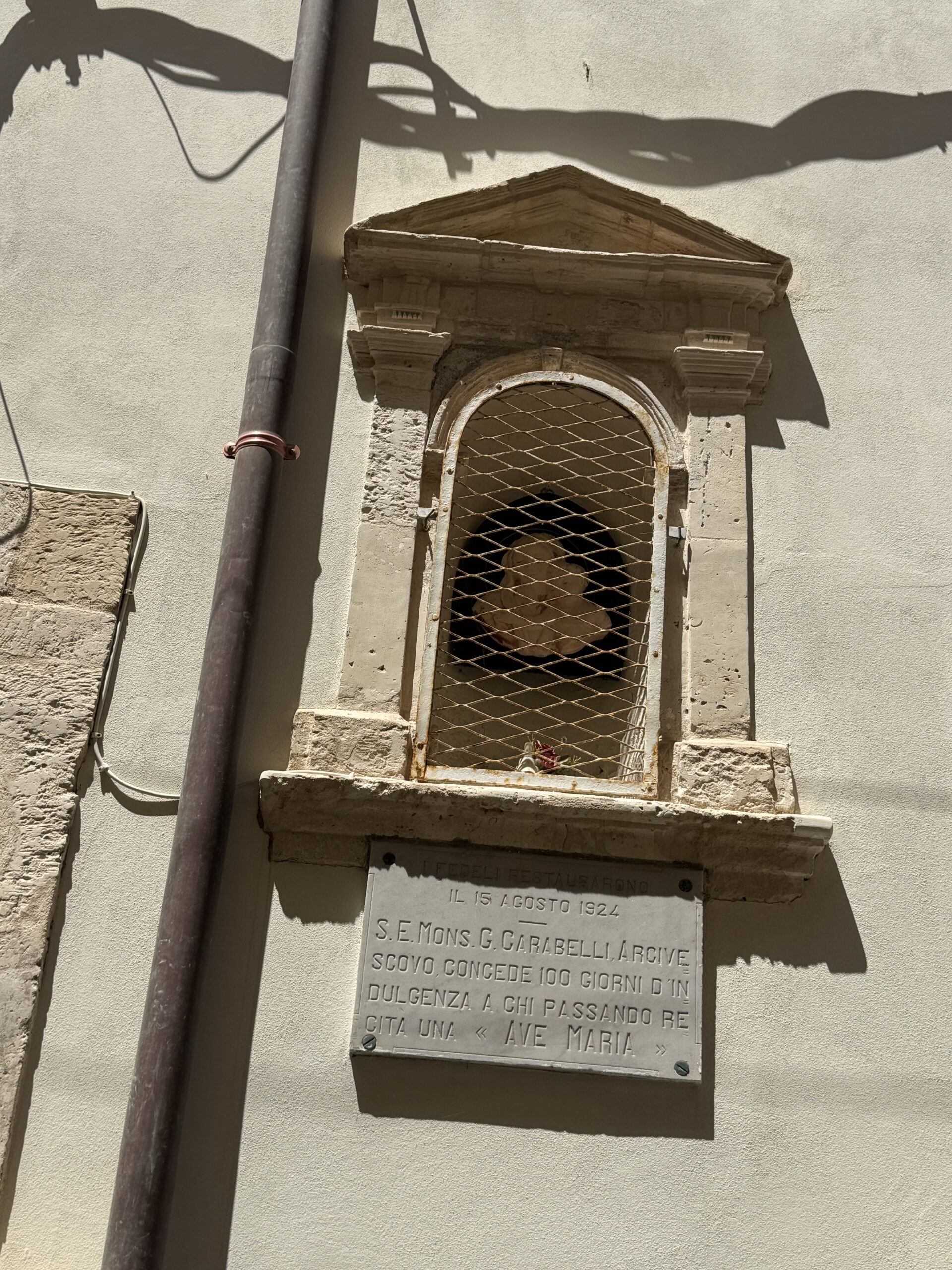 Edicola votiva a muro con timpano triangolare, nicchia ad arco chiusa da grata romboidale e lapide marmorea sottostante.