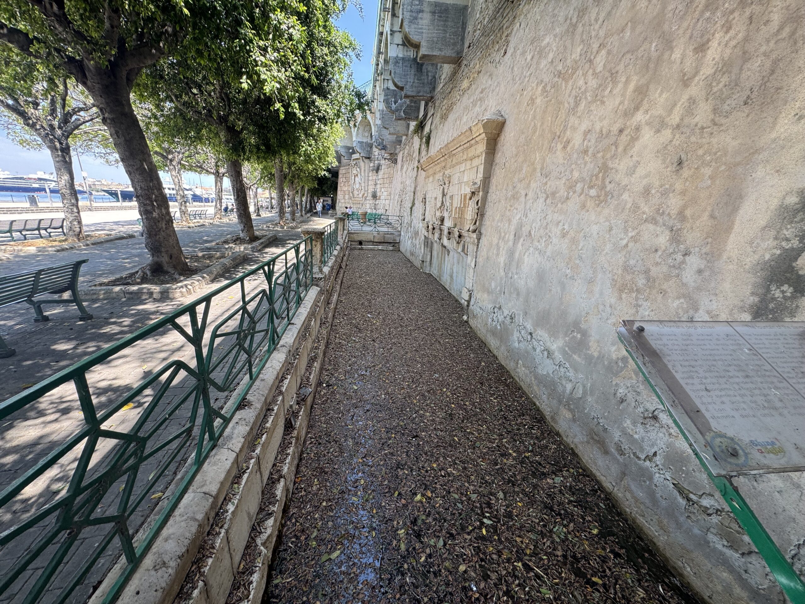 La Fontana degli Schiavi, lungo prospetto in pietra in un recinto ribassato lungo il Foro Italico di Ortigia, con la passeggiata alberata e il mare sullo sfondo.
