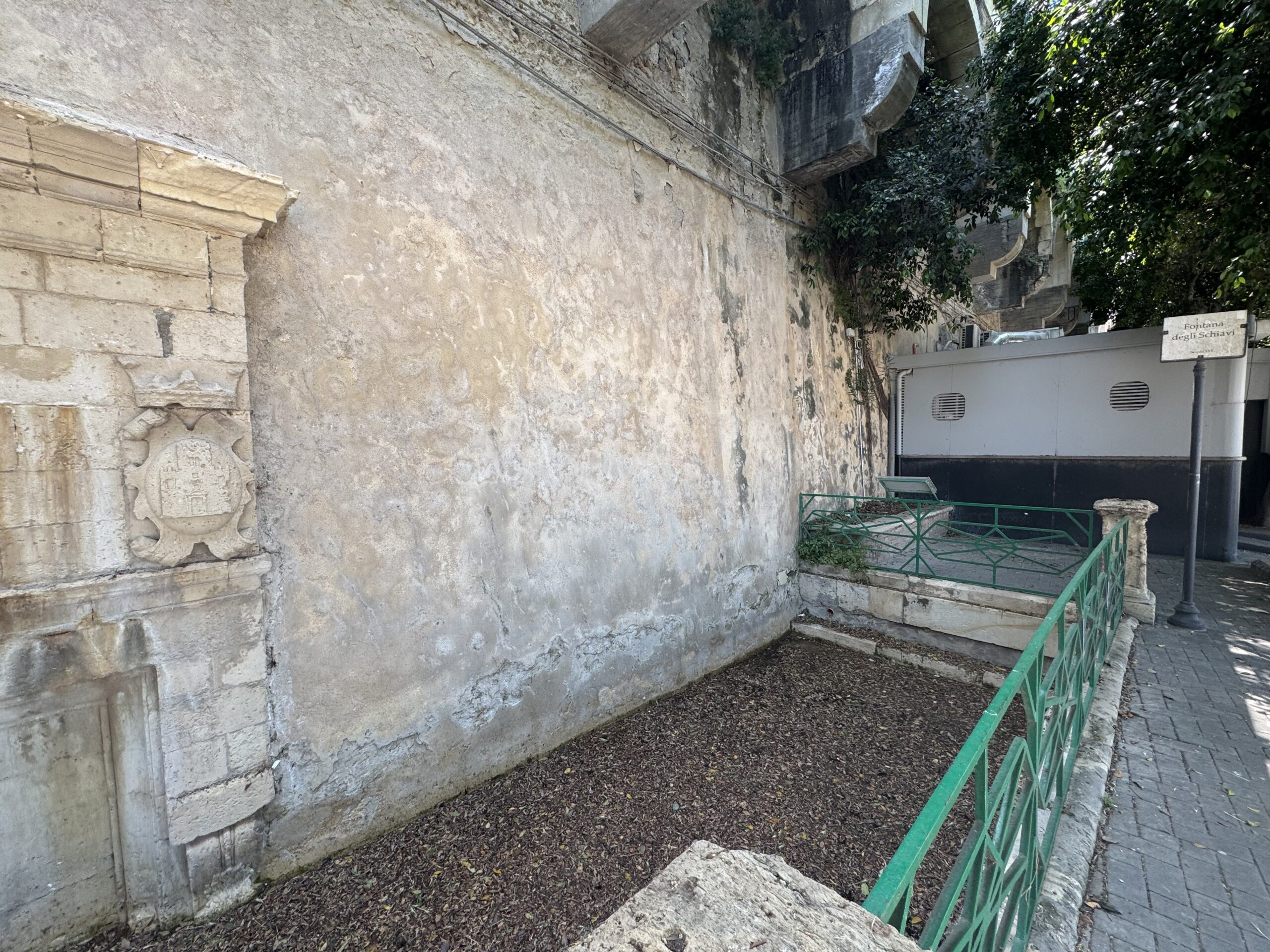 L'estremità scolpita del prospetto della Fontana degli Schiavi, con uno stemma entro cartella, addossata alla muraglia della Marina di Ortigia.