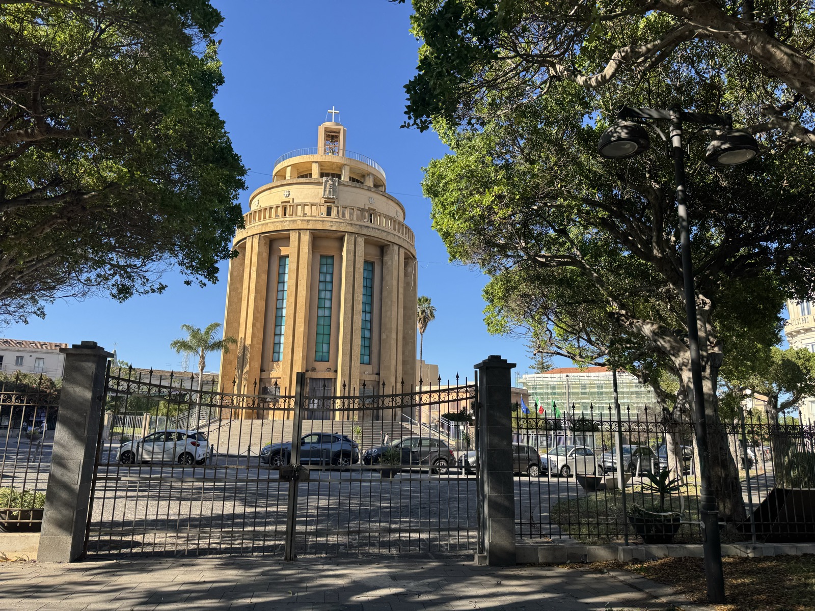 Vista a tre quarti del Pantheon dei Caduti vista attraverso la cancellata perimetrale, con auto parcheggiate e la cupola illuminata dal sole.