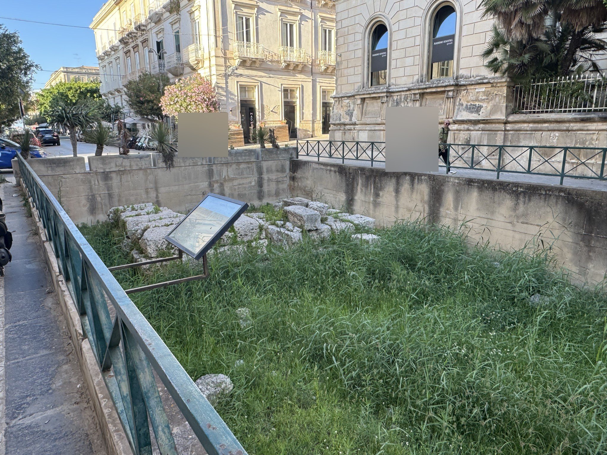 Panoramica dell'area archeologica recintata con il pannello informativo in primo piano e i palazzi di Ortigia sullo sfondo.