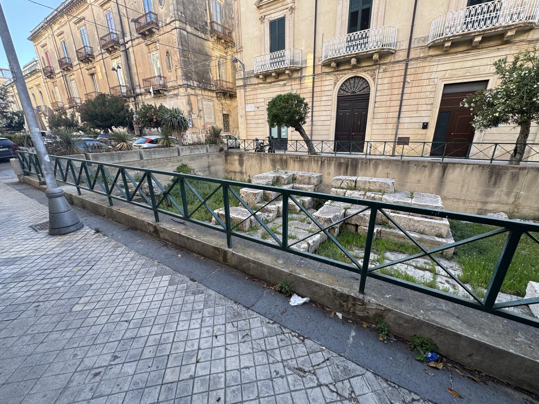 Recinzione perimetrale verde dell'area archeologica con il marciapiede pedonale e i palazzi residenziali di Ortigia di fronte.
