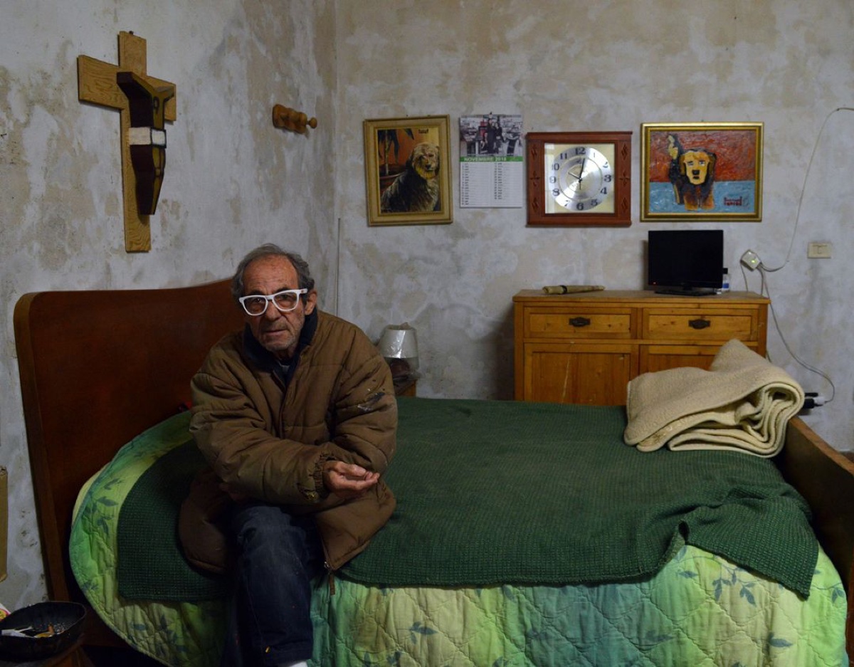 Ritratto del pittore Salvatore Accolla seduto sul letto della sua casa di Ortigia con due delle sue opere appese al muro, crocifisso ligneo e calendario di novembre 2018