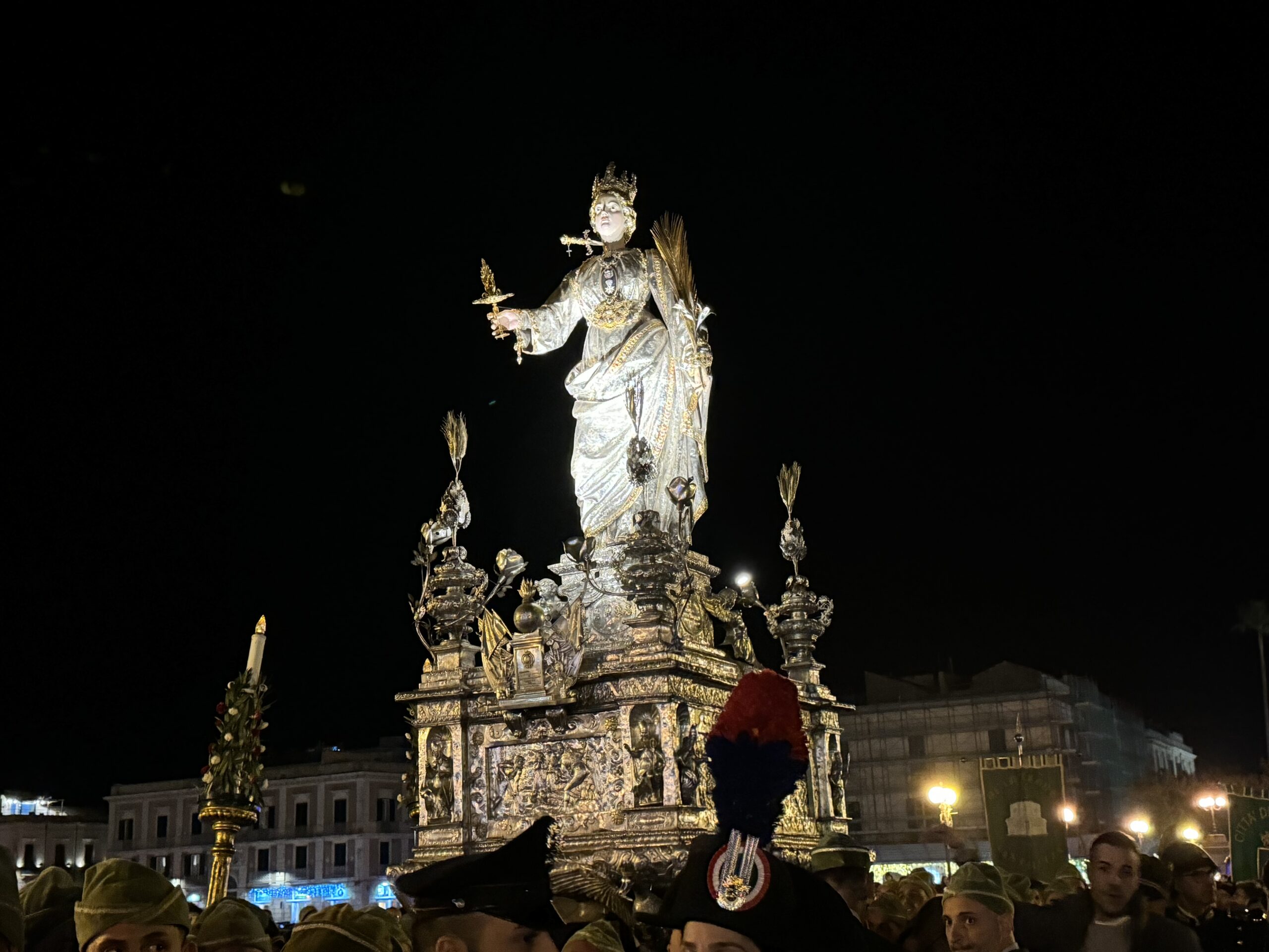 Simulacro argenteo di Santa Lucia illuminato di notte durante la processione del 13 dicembre 2023, folla con berretti verdi e Carabinieri