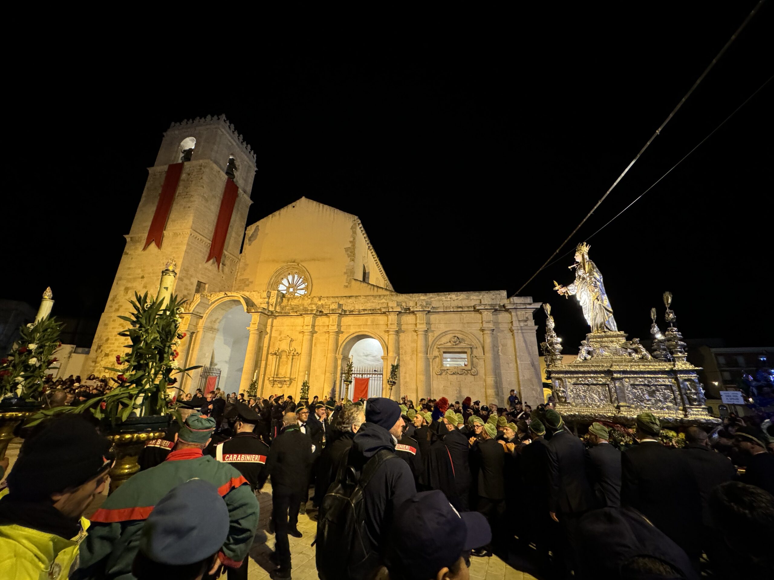 Simulacro argenteo di Santa Lucia di notte davanti a edificio storico durante la processione del 13 dicembre 2023, folla con berretti verdi e Carabinieri