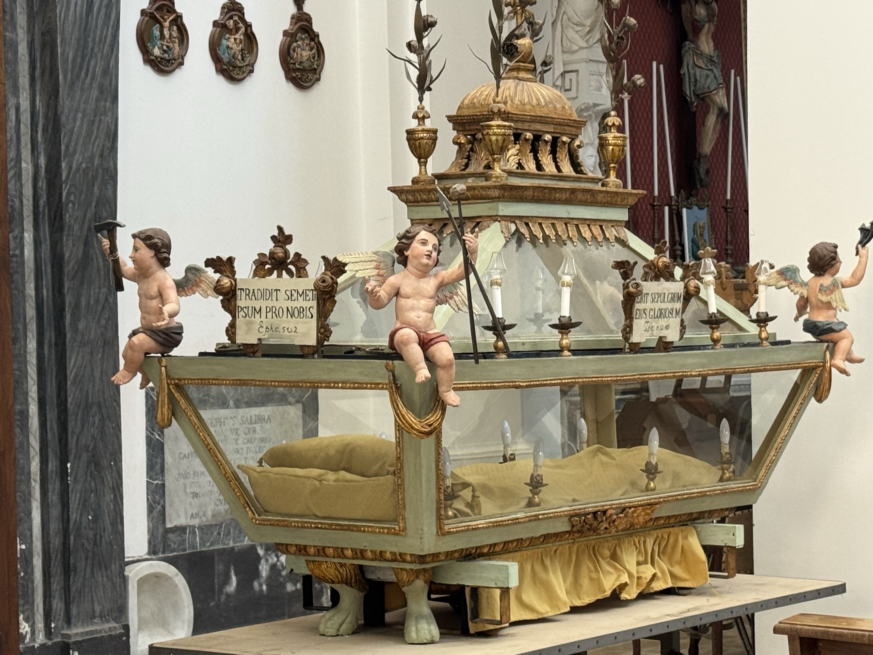 Urna processionale lignea verde e oro con teca di vetro, sormontata da tre putti alati che reggono i simboli della Passione, con cartigli e iscrizioni latine.