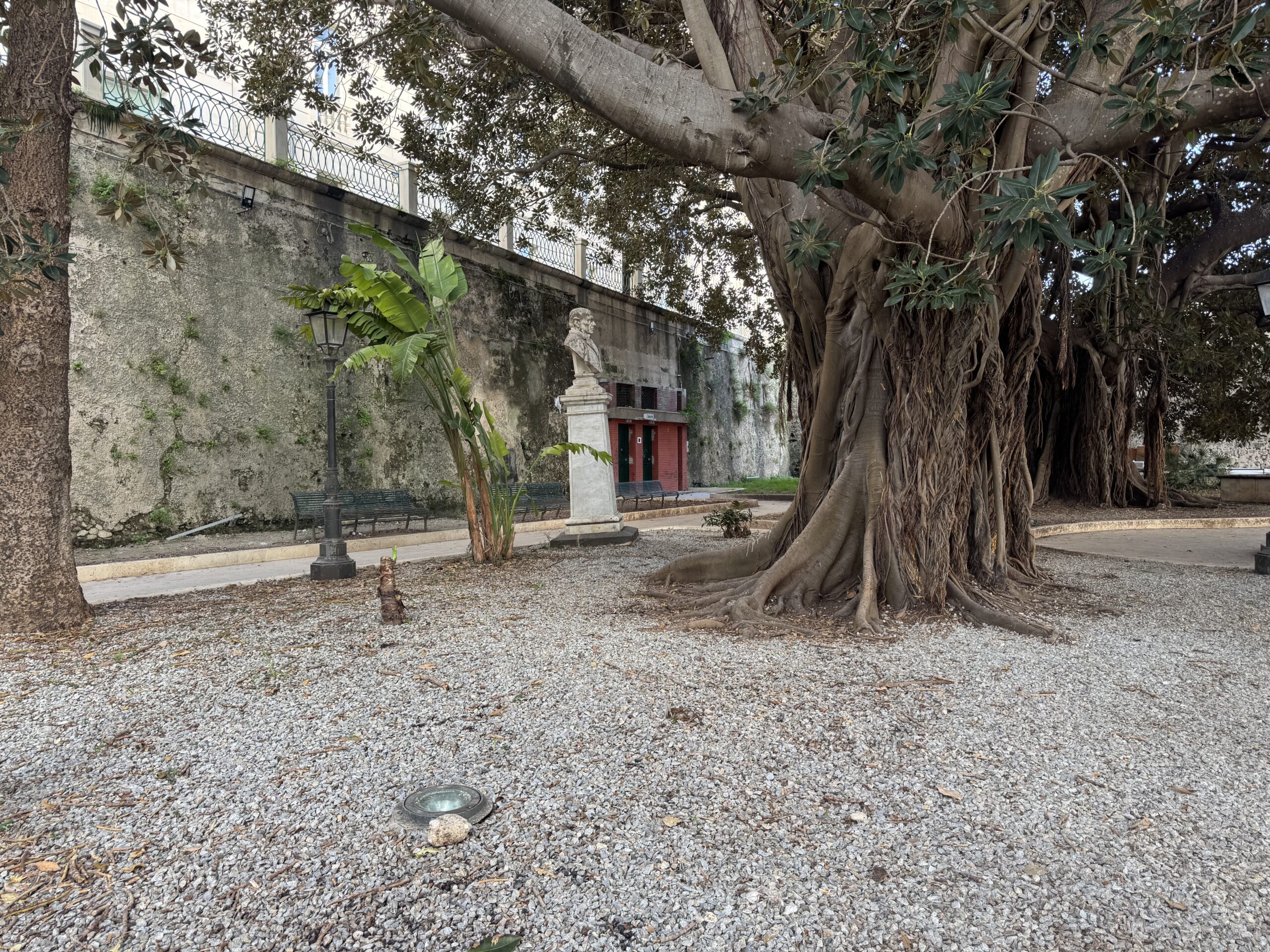 Vista ampia della Villetta Aretusa con un ficus monumentale in primo piano, lampione ottocentesco, panchine in ghisa e il busto a Campisi sullo sfondo.