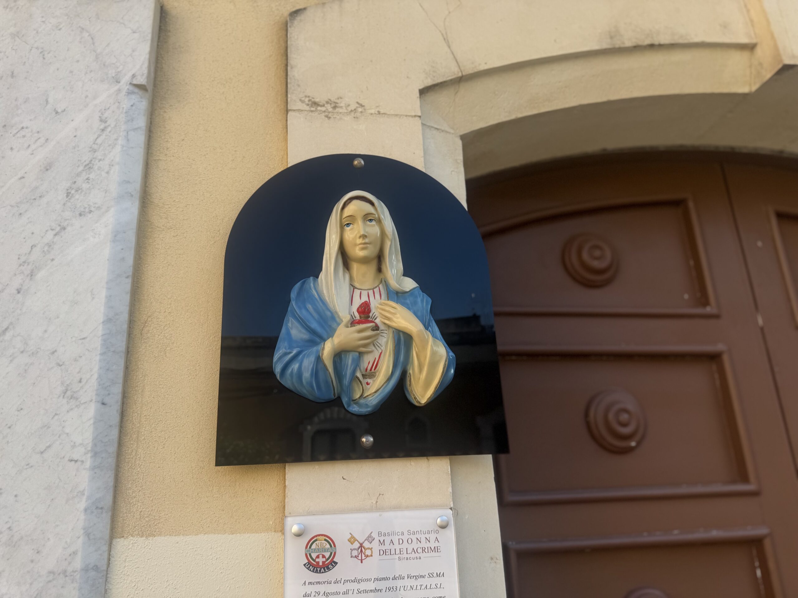 Bassorilievo policromo della Madonna con manto azzurro e Cuore Immacolato sul petto, montato su uno scudo blu navy accanto al portone in legno marrone