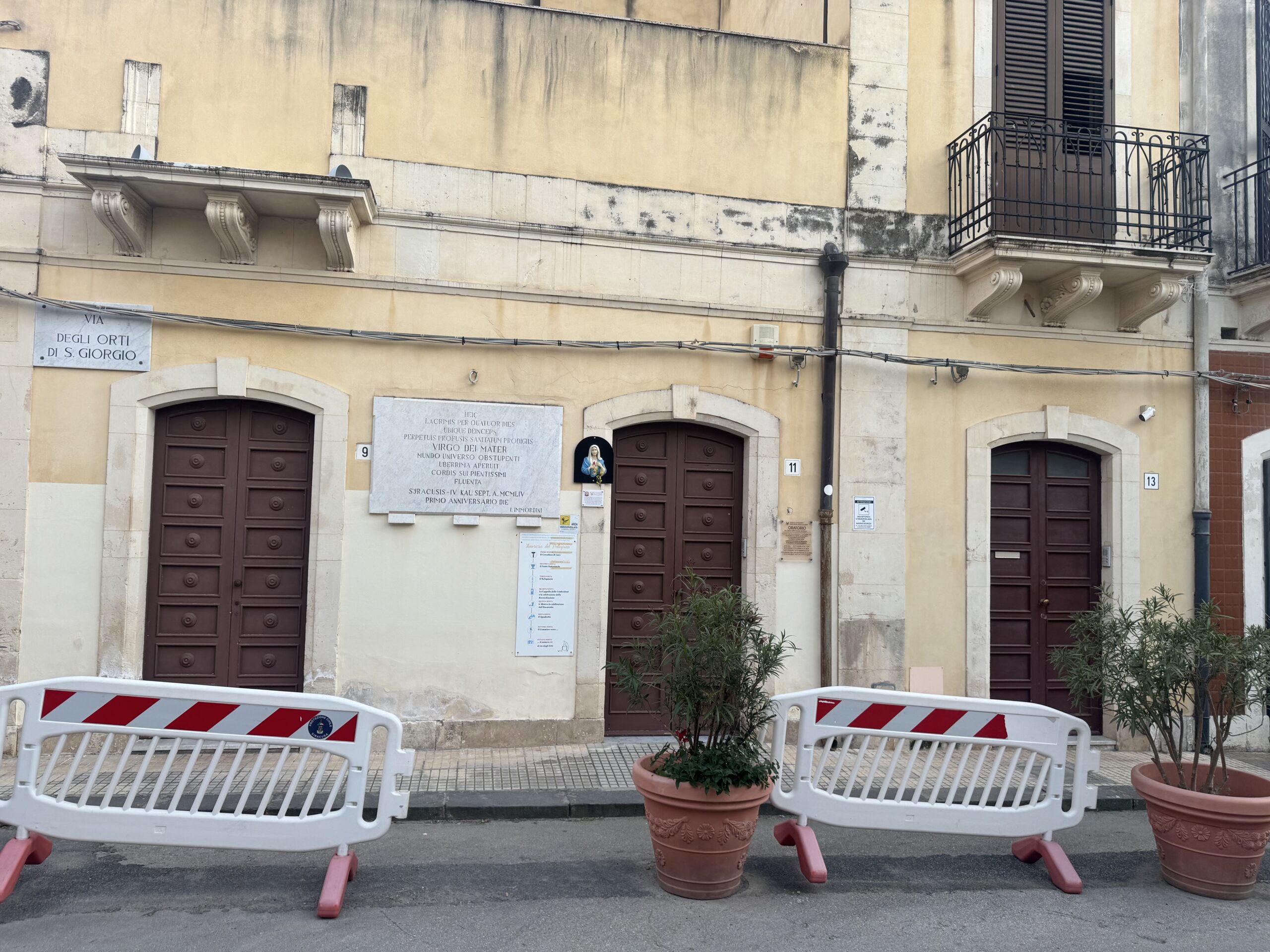 Facciata in intonaco ocra della Casa del Pianto a Siracusa, con tre portoni in legno marrone, la targa stradale di via degli Orti di San Giorgio, i civici 9 e 11, la lapide marmorea e il bassorilievo della Madonna