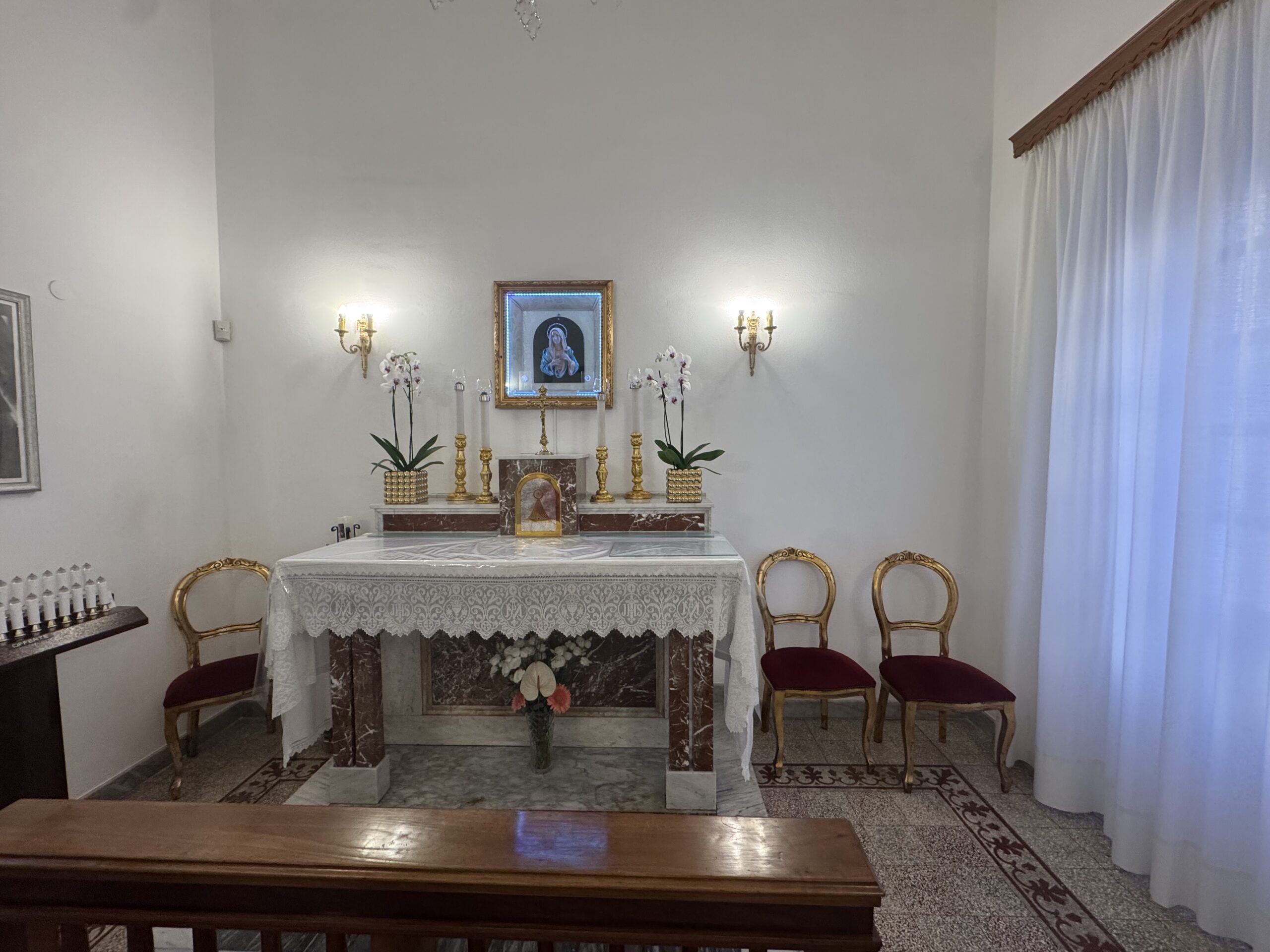 Piccola sala a pareti bianche con altare in marmo rosso e tovaglia di pizzo, quadretto del Cuore Immacolato retroilluminato sopra l'altare, due appliques dorate, orchidee bianche, sedie dorate e una panca lignea