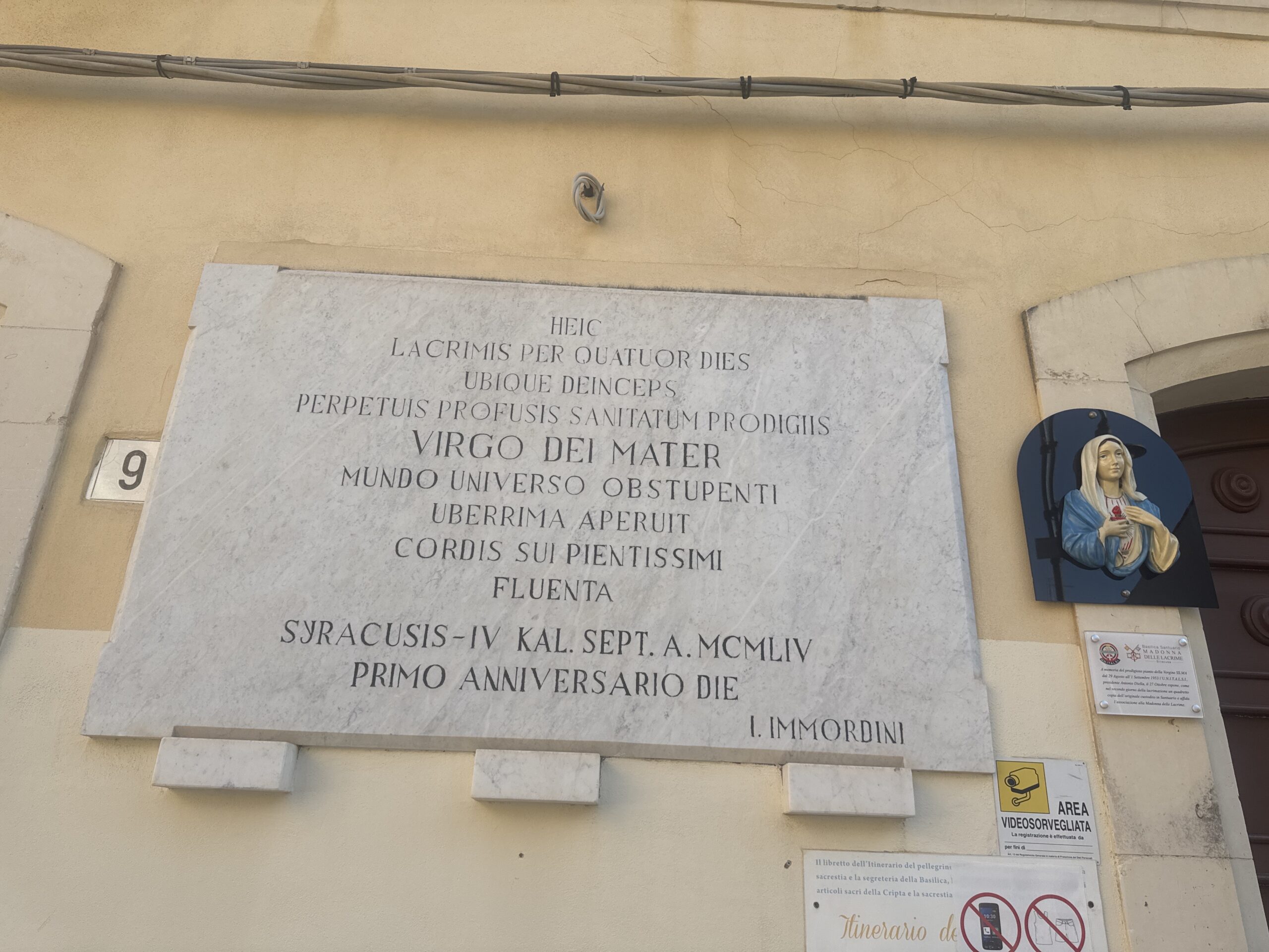Grande lapide in marmo bianco murata sulla facciata ocra, con lunga iscrizione latina a lettere capitali e, a destra, il bassorilievo della Madonna accanto al portone